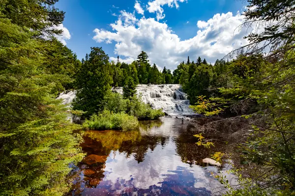 Waterfall in a scenic forest setting, with a river flowing through lush greenery under a bright, cloudy sky